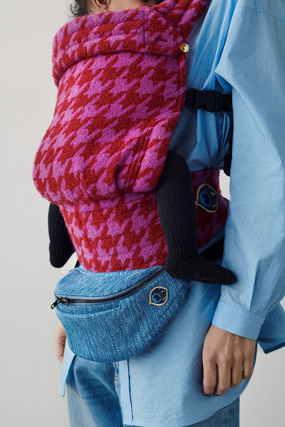 A close-up of a model wearing a red and pink tweed cashmere and silk baby carrier and 
denim belt bag